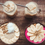 Zwei Smoothie Bowls in Kokosnuss Schalen mit Banane, Granola und Kokos, dazu Iced Coffee im Glas