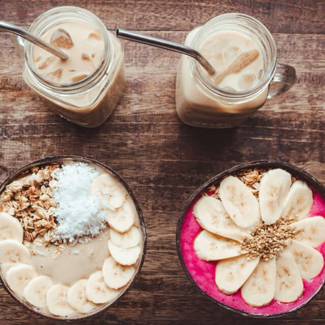 Zwei Smoothie Bowls in Kokosnuss Schalen mit Banane, Granola und Kokos, dazu Iced Coffee im Glas