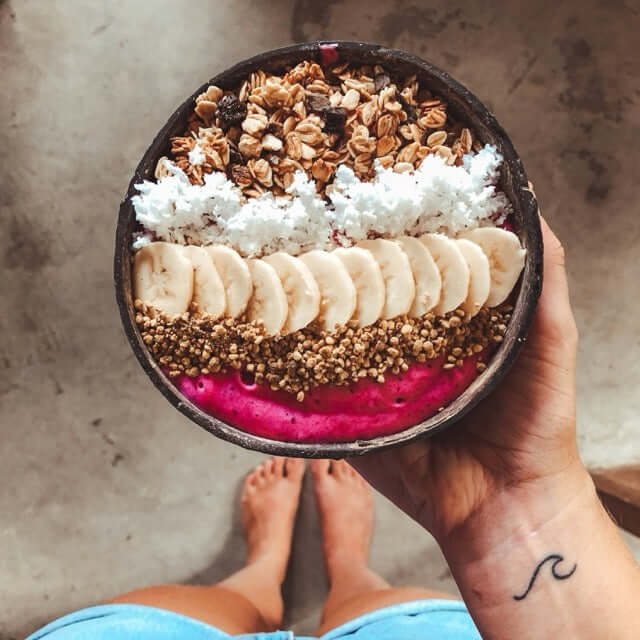 Hand hält pinke Smoothie Bowl in Kokosnuss Schale, belegt mit Banane, Granola und Kokosflocken